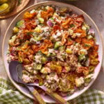 A bowl of salad with crispy golden pieces, chopped cucumbers, red onions, pickles, and fresh dill, served with wooden utensils on a green checkered cloth. A plate of pickles and a bowl of white sauce are nearby.