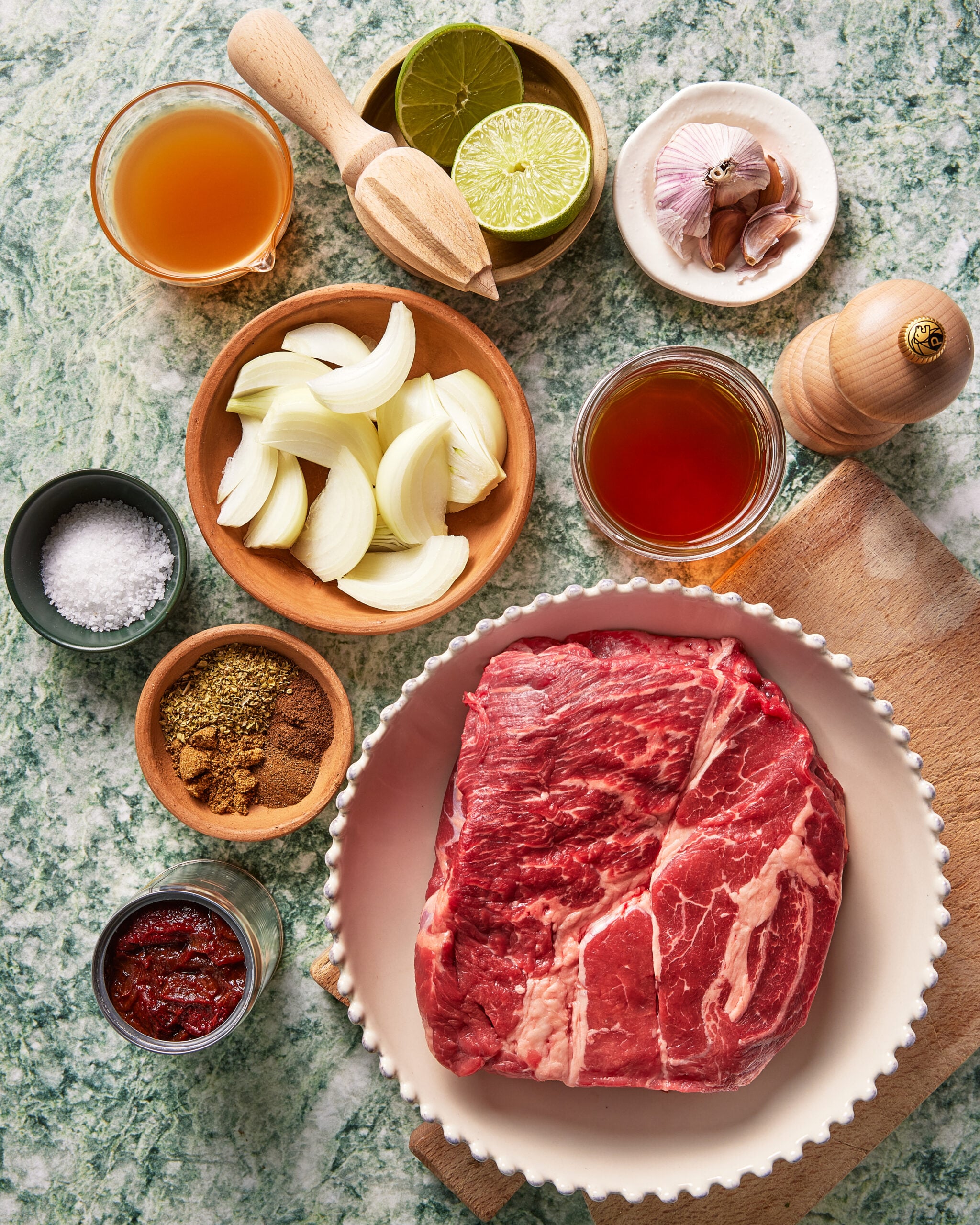 Raw beef in a bowl on a cutting board surrounded by sliced onions, garlic, halved limes, spices, salt, pepper, broth, and tomato paste, all arranged on a green marble countertop.