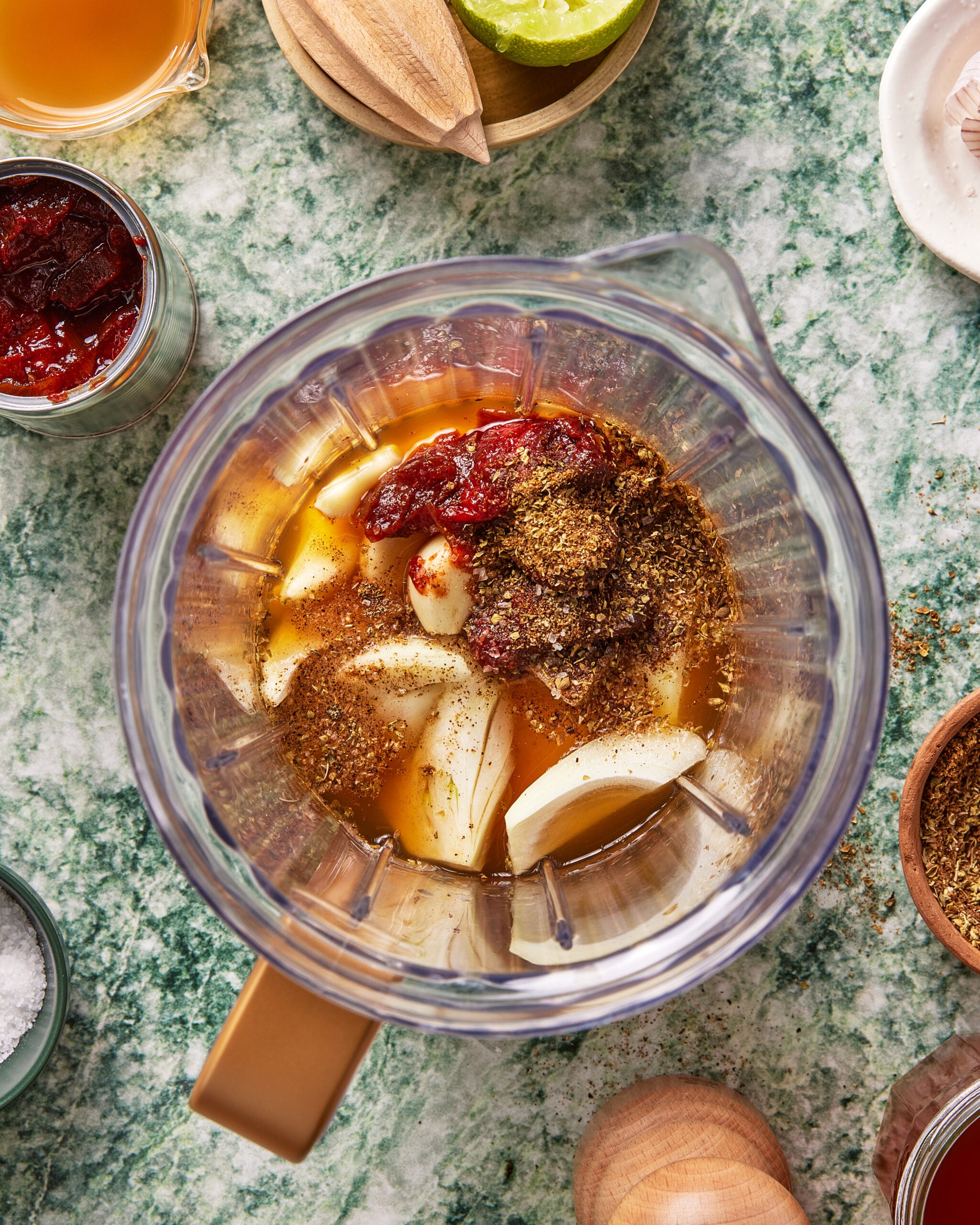 Overhead view of a blender containing onion slices, garlic cloves, dried spices, ketchup, and a liquid ingredient, placed on a green marble countertop surrounded by seasonings and recipe ingredients.