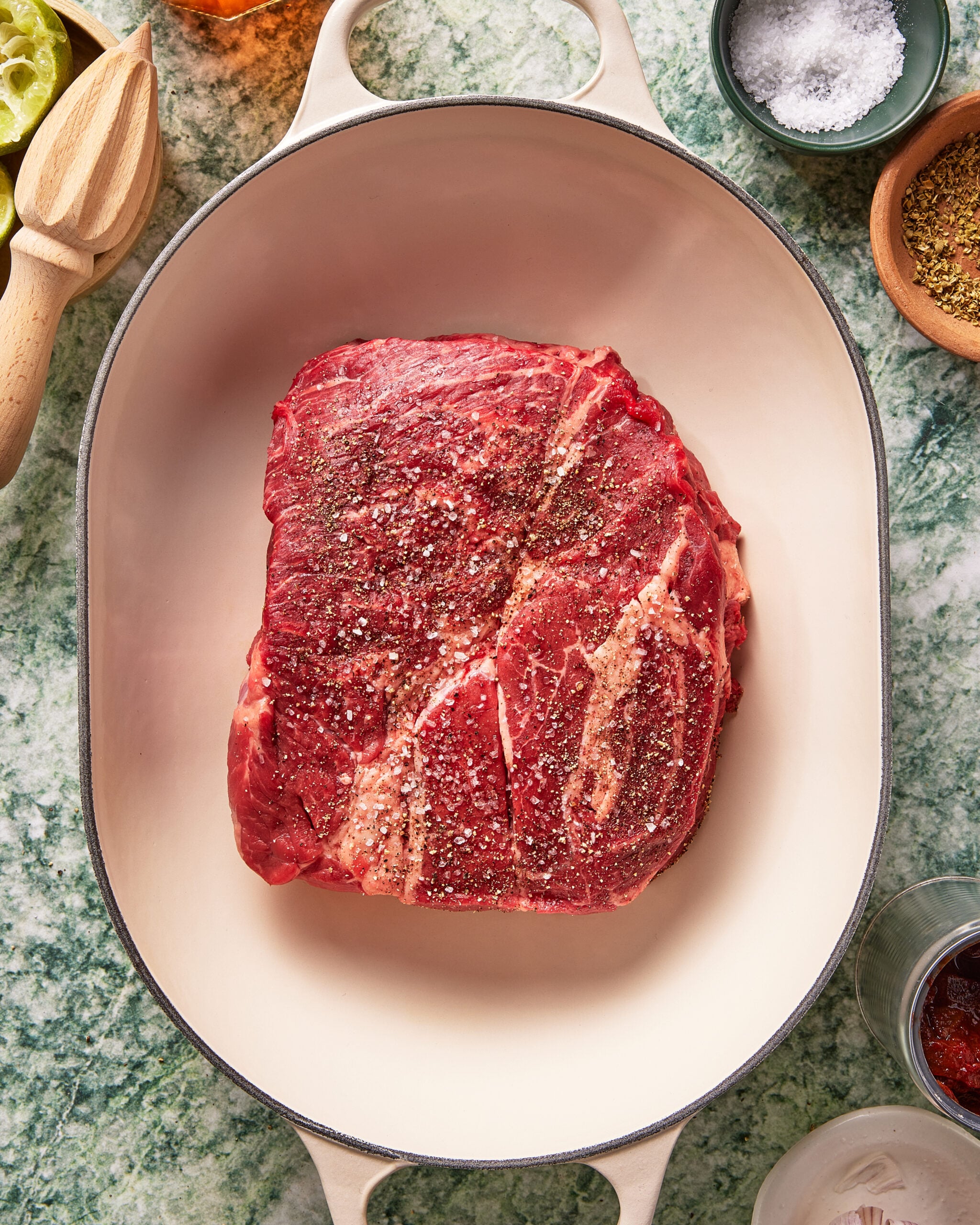 A raw, seasoned beef steak sits in a large, oval white Dutch oven, surrounded by small bowls of spices and salt on a green marble countertop.