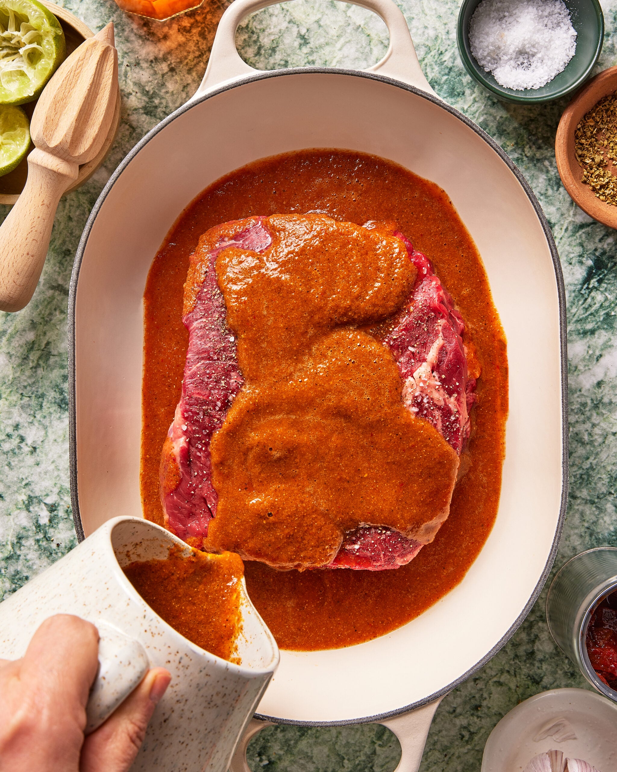 A hand pours reddish marinade over a raw beef roast in an oval white baking dish on a green marble counter, surrounded by bowls of salt, spices, and lime halves.