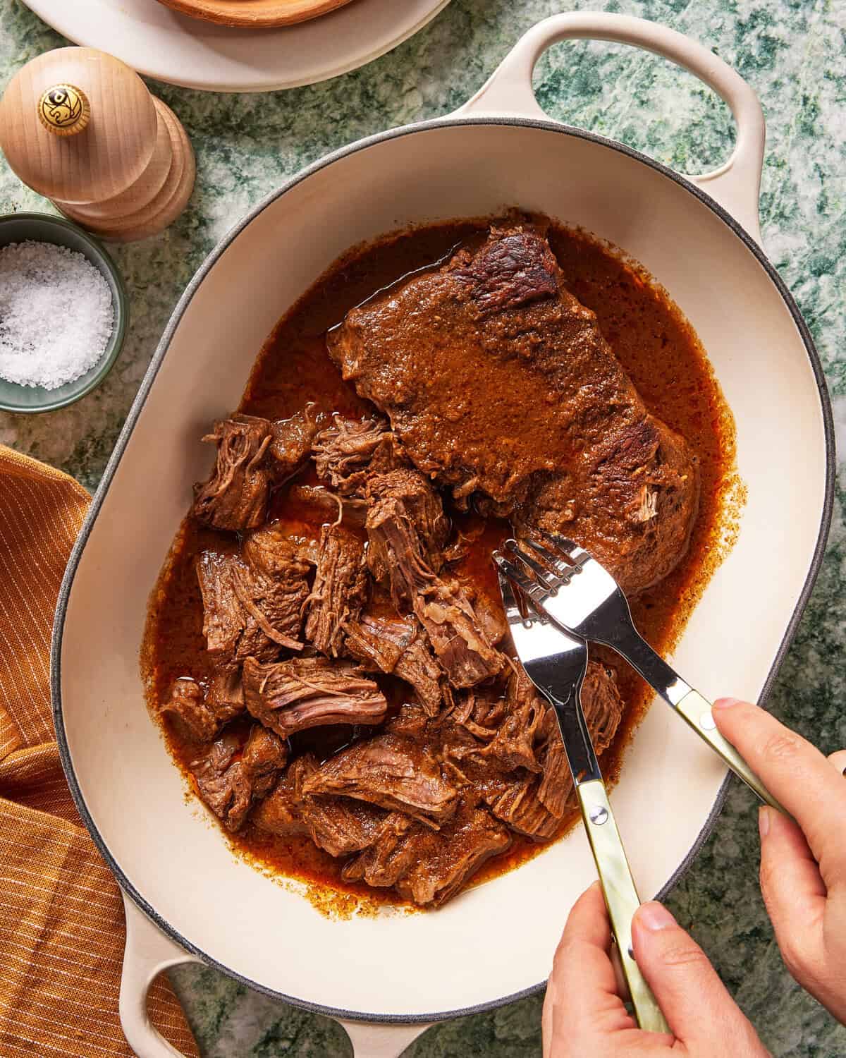 A person uses two forks to shred tender cooked beef in a white oval dish filled with rich, brown sauce. A bowl of salt, pepper mill, and orange napkin are nearby on a green countertop.
