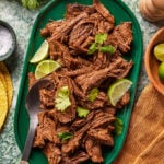 Shredded beef barbacoa garnished with cilantro and lime wedges on a green platter, with a serving spoon beside the meat. Tortillas, salt, and more cilantro visible nearby on a marble surface.