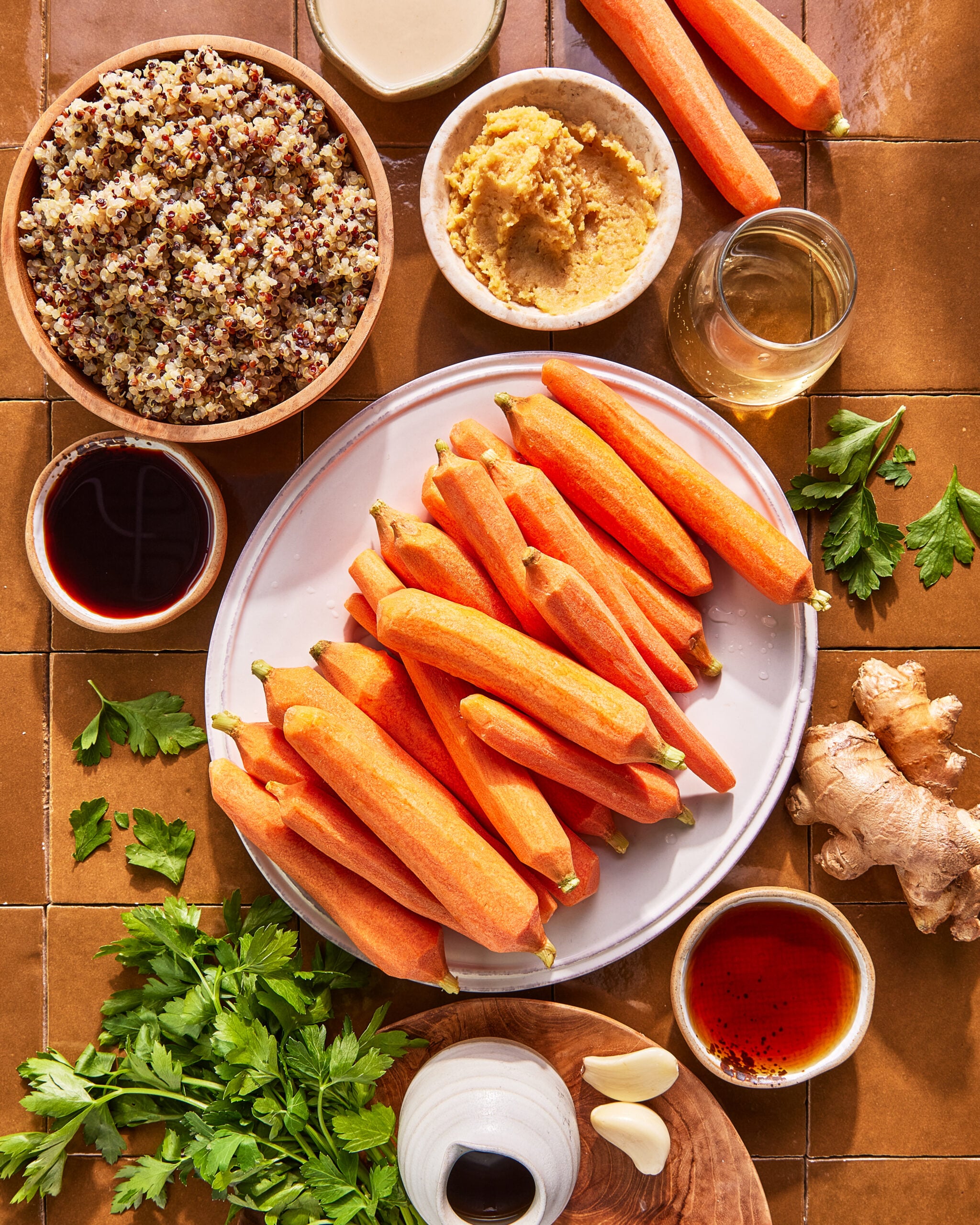 Overhead view of ingredients on a brown tiled surface, including peeled carrots on a plate, a bowl of quinoa, a bowl of hummus, jars of oil and vinegar, fresh parsley, ginger, garlic, and small bowls of sauces.