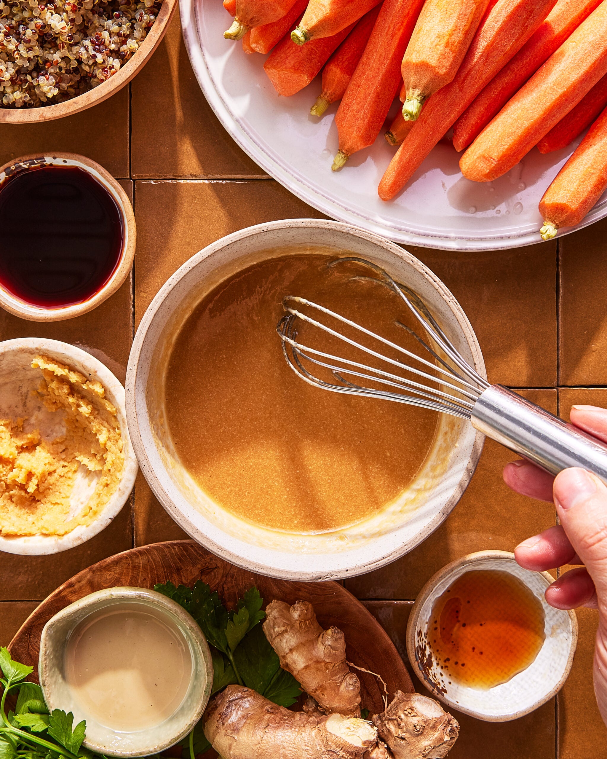 A hand whisking a creamy sauce in a bowl, surrounded by fresh carrots, grated ginger, parsley, ginger root, quinoa, tamari, tahini, and a small bowl of oil on a brown tiled surface.