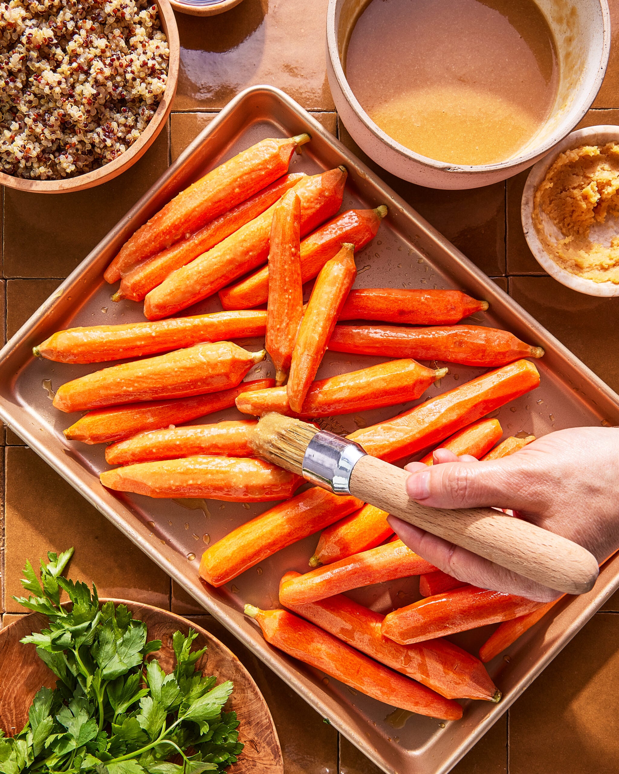 A hand uses a brush to coat peeled carrot sticks on a baking sheet. Surrounding the tray are bowls containing quinoa, sauce, hummus, and fresh parsley on a tiled surface.