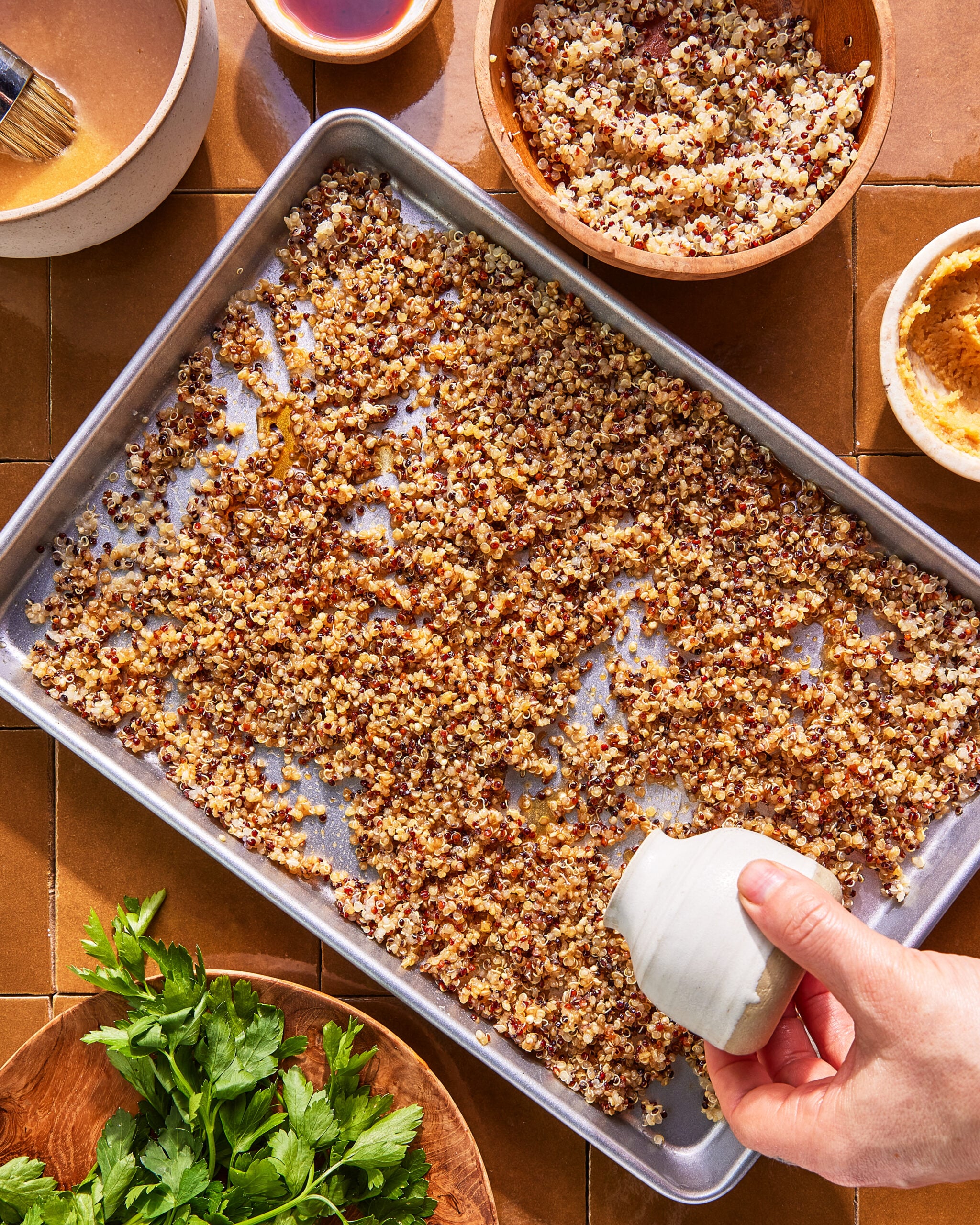 A hand pours oil from a small white container over cooked quinoa spread on a baking sheet. Bowls of quinoa, herbs, and dressing are on a brown tiled surface nearby.