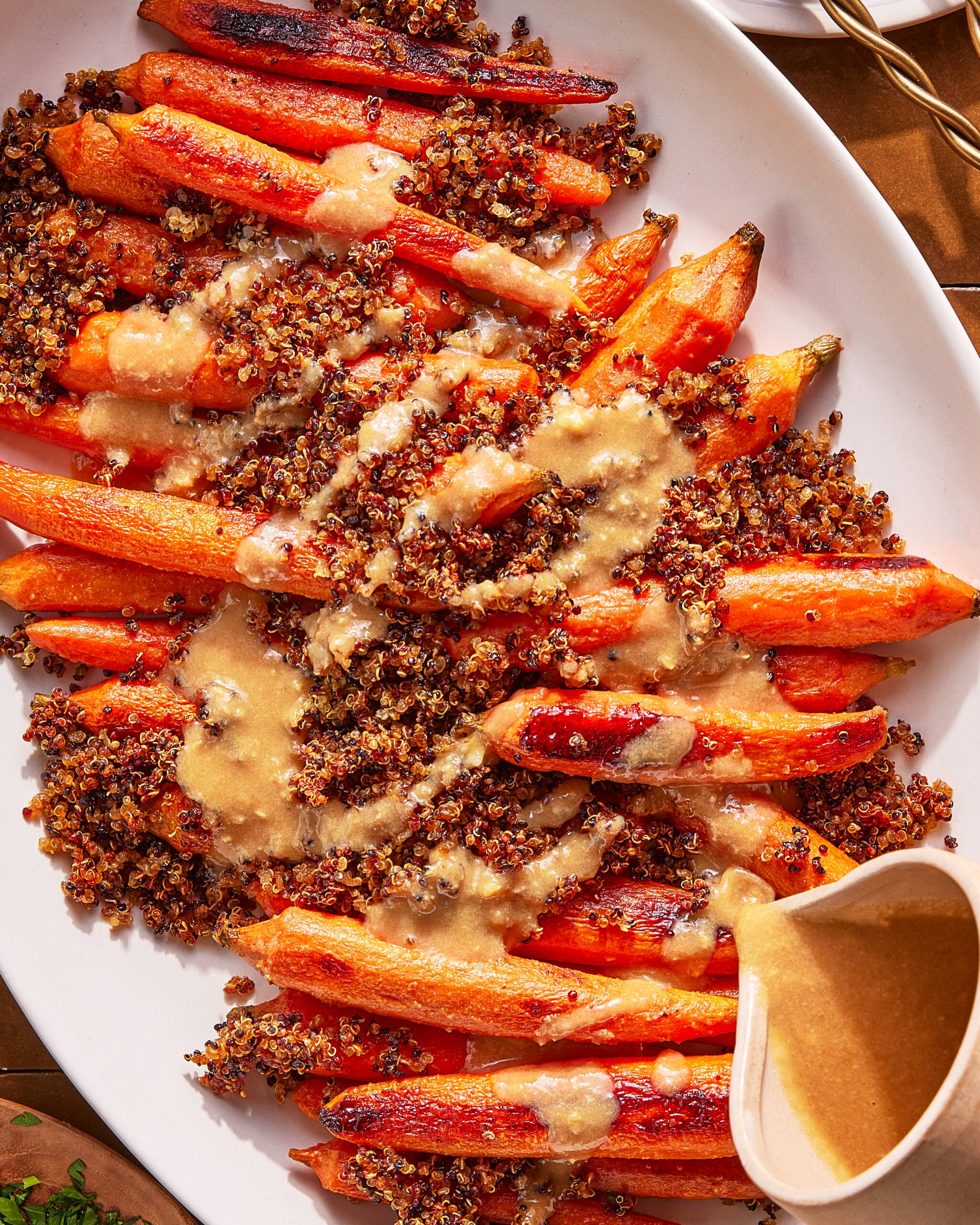 Roasted carrots are topped with a layer of cooked quinoa and drizzled with a creamy, light-brown sauce, served on a white oval platter with extra sauce in a small pitcher on the side.