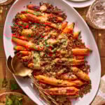A white oval platter with roasted carrots on a bed of quinoa, drizzled with a creamy dressing and garnished with chopped herbs. A gold serving spoon rests on the platter, with plates and glasses nearby.