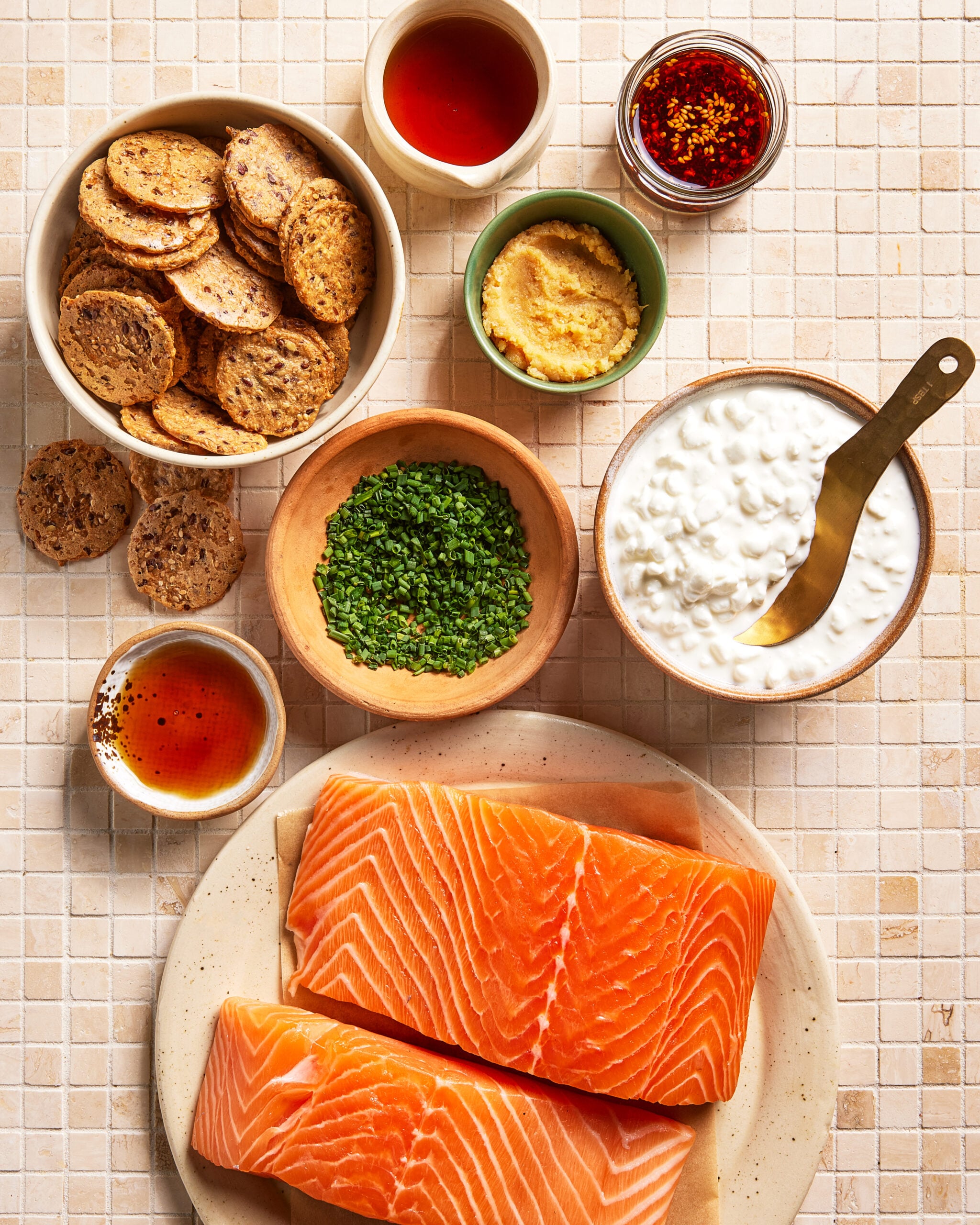 Two raw salmon fillets on a plate surrounded by bowls of crackers, cottage cheese, chopped chives, Dijon mustard, honey, soy sauce, and red pepper flakes, all arranged on a tiled surface.