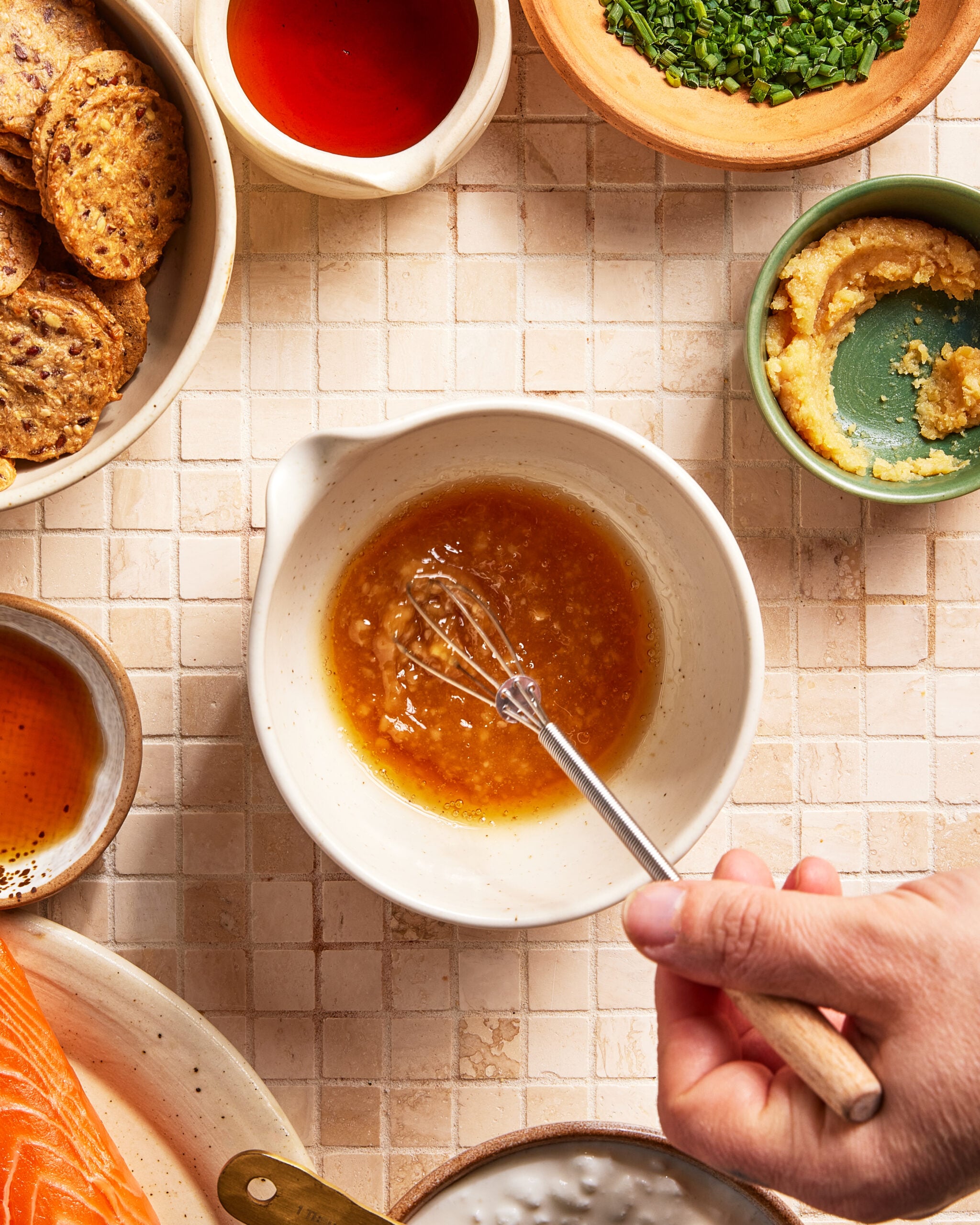A hand whisks a brown liquid in a white bowl on a tiled surface, surrounded by crackers, chopped herbs, salmon, a red sauce, and a bowl of creamy mixture.
