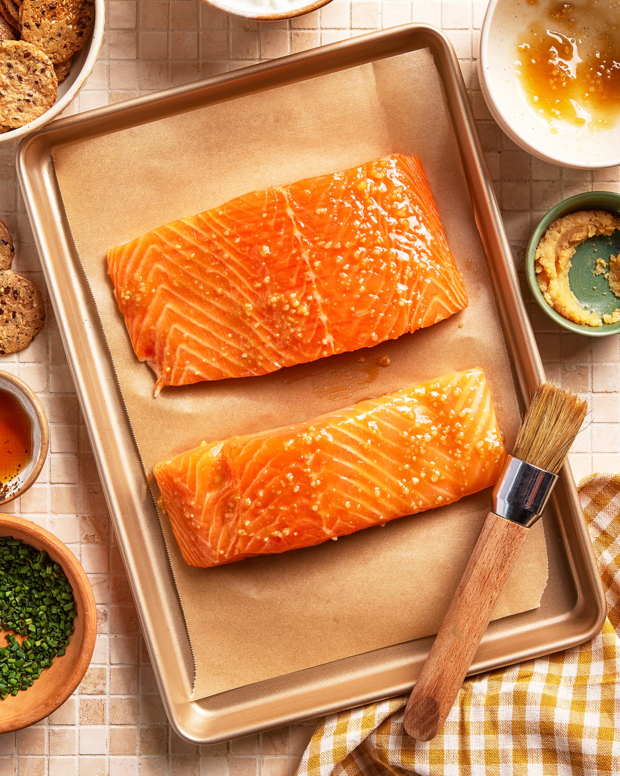 Two raw salmon fillets brushed with glaze and seasoned with sesame seeds on a parchment-lined baking tray, surrounded by small bowls of sauces, crackers, chives, and a basting brush.