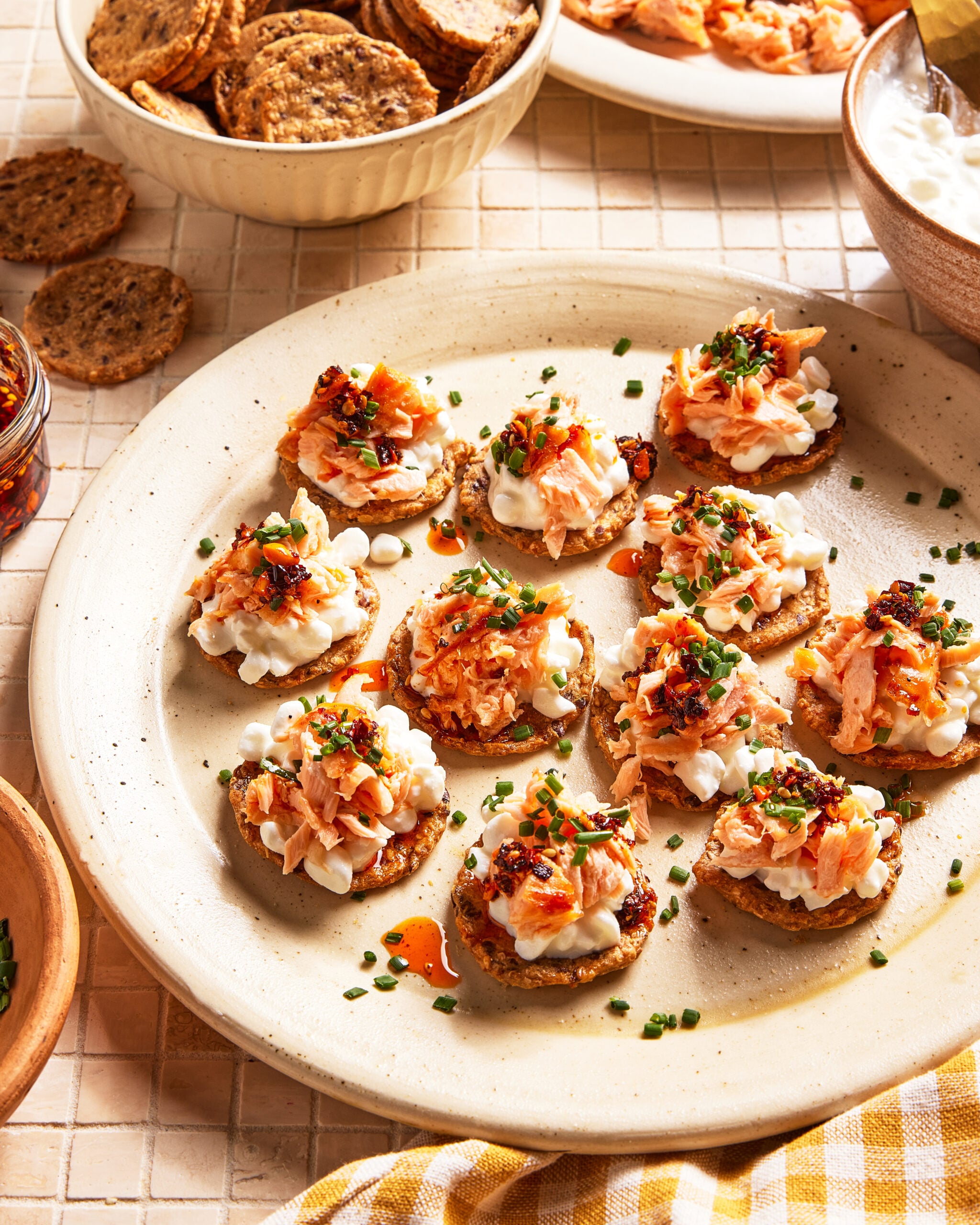 A plate of bite-sized crackers topped with a creamy spread, shredded salmon, chopped chives, and a drizzle of chili oil. Bowls of dip and extra crackers are in the background on a tiled surface.