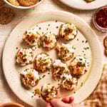 A hand picking up a round cracker topped with a creamy spread, chives, and seasoning from a plate filled with similar crackers. Surrounding the plate are crackers, a bowl of chopped chives, and a dish of chili oil.