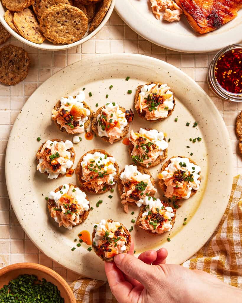 A hand picking up a round cracker topped with a creamy spread, chives, and seasoning from a plate filled with similar crackers. Surrounding the plate are crackers, a bowl of chopped chives, and a dish of chili oil.