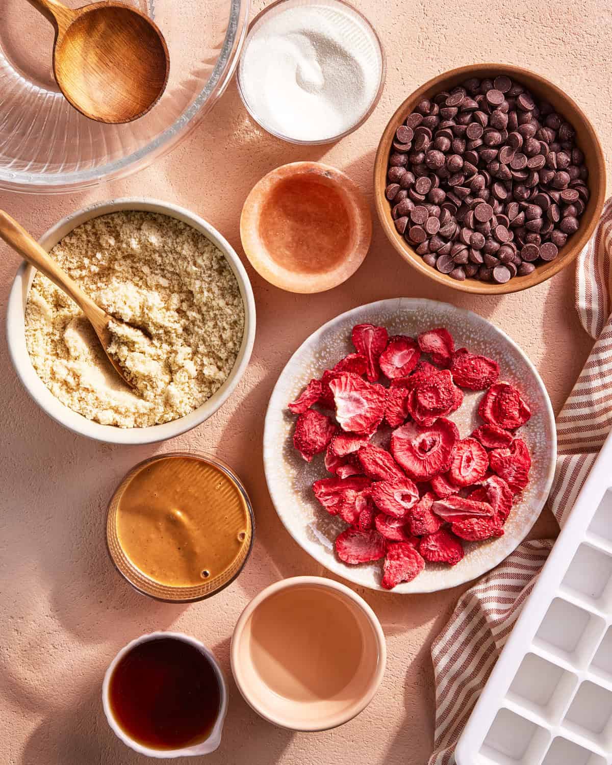 A flat lay of baking ingredients on a pink surface, including chocolate chips, almond flour, freeze-dried strawberries, sugar, syrup, and various wooden and glass bowls, with an ice cube tray and striped cloth nearby.