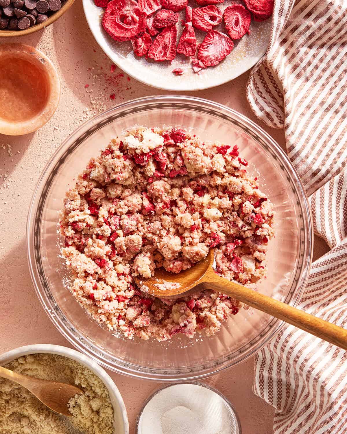 A glass bowl filled with a crumbly strawberry mixture being stirred with a wooden spoon. Surrounding the bowl are plates and bowls with strawberries, chocolate chips, sugar, and a striped cloth napkin.