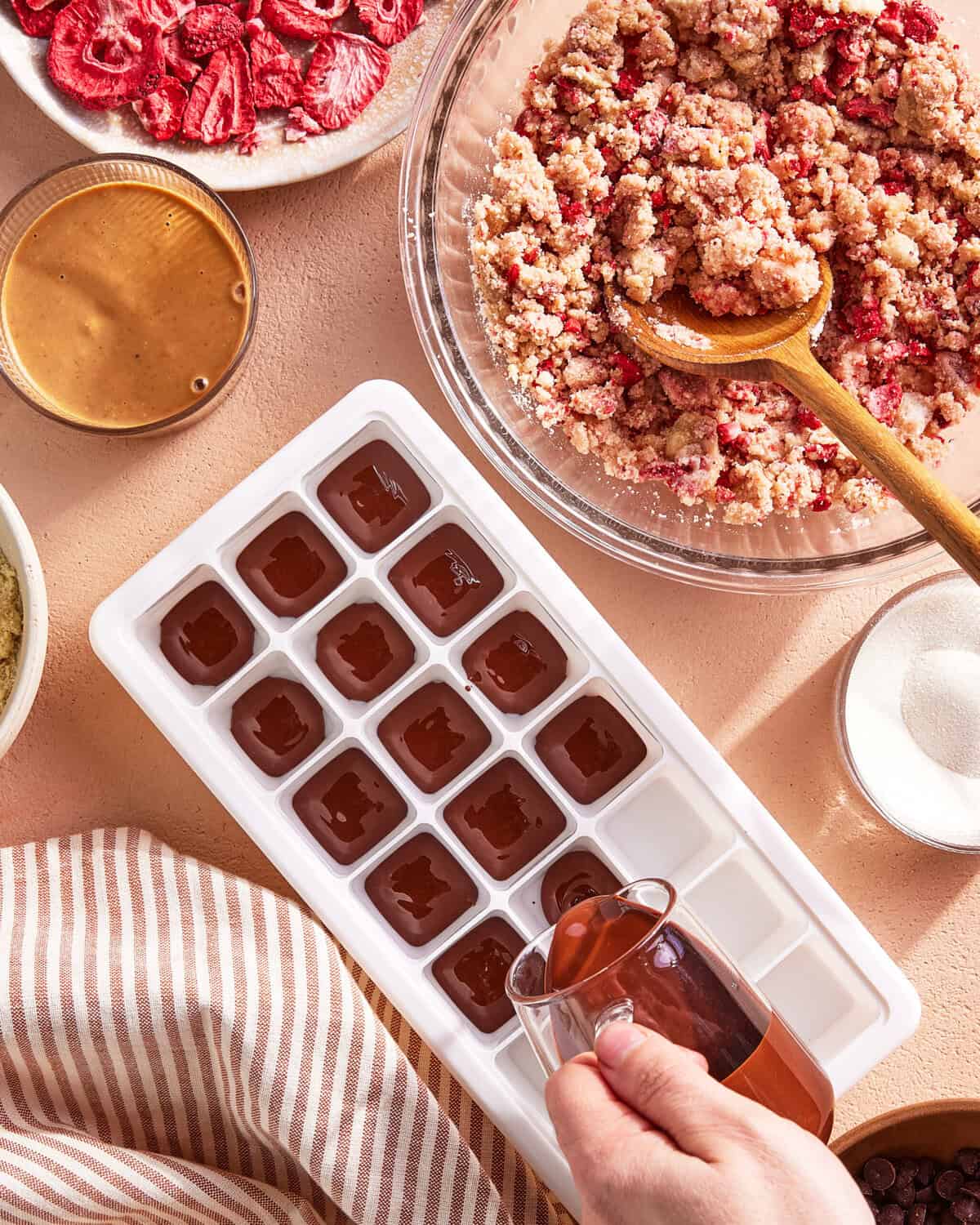 A hand pours chocolate mixture into an ice cube tray. Nearby are a bowl of crumbly strawberry mixture, a plate of dried strawberries, a bowl of peanut butter, and a jar of sugar on a beige surface.
