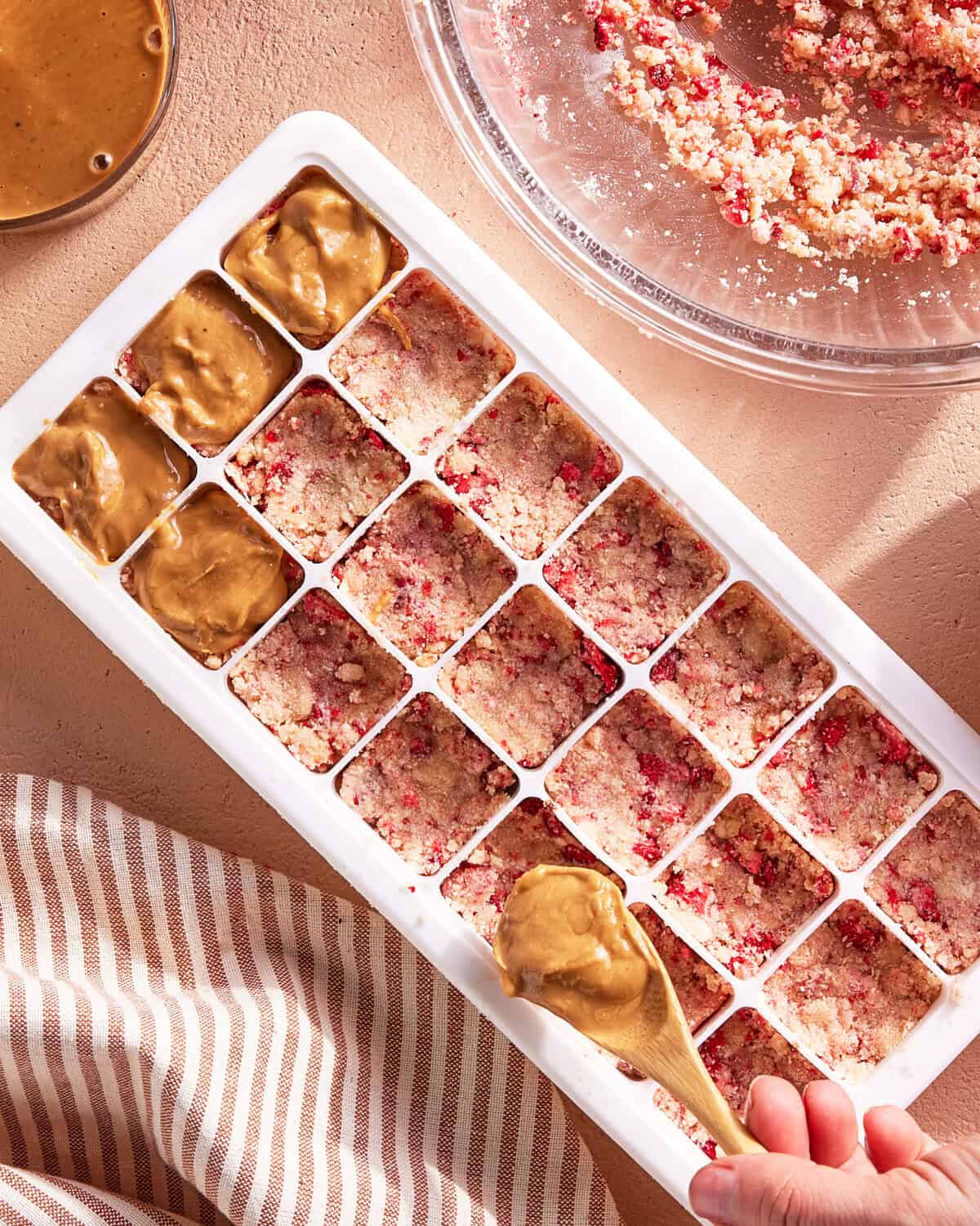 An ice cube tray filled with a crumbly pink and white mixture, partially topped with smooth brown peanut butter. A hand holds a spoon, spreading peanut butter over the mixture. A striped cloth and mixing bowl are nearby.