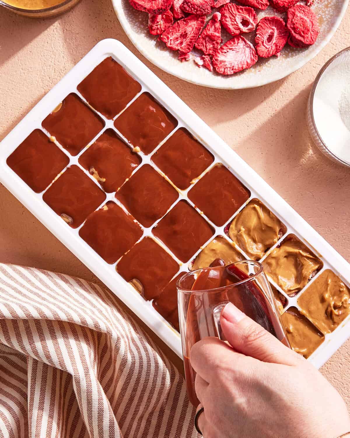 A hand pours melted chocolate into an ice cube tray, filling compartments partially occupied by peanut butter. Nearby are bowls of freeze-dried strawberries and sugar, and a striped towel rests on the counter.