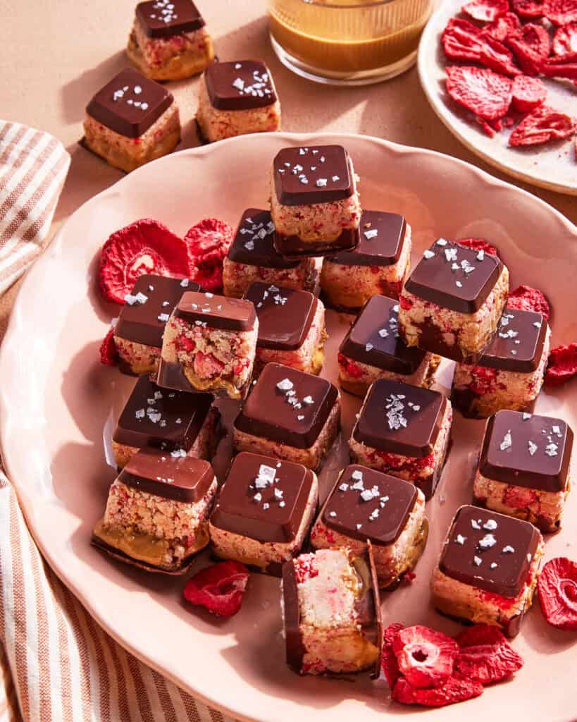 A plate of square chocolate-covered treats with a nutty and fruity base, topped with sea salt, surrounded by dried strawberry slices. Some treats are stacked, showing their layered interior.
