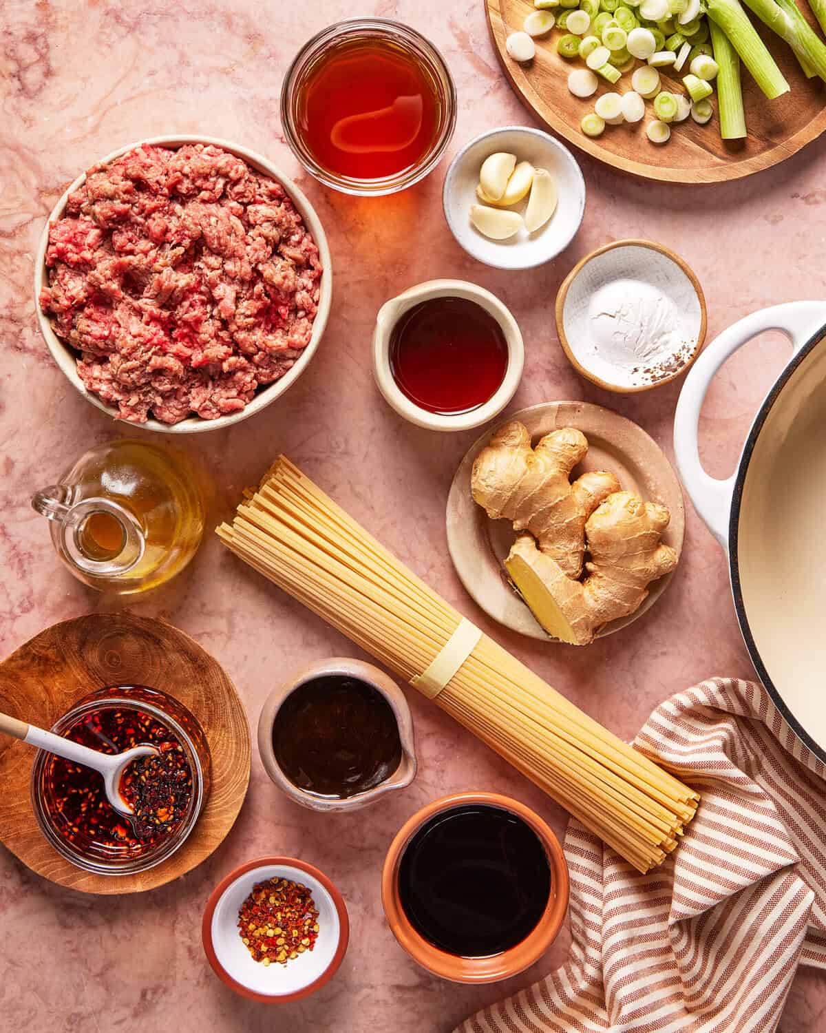 Overhead view of various ingredients on a counter, including ground meat, dried noodles, ginger, garlic, green onions, chili flakes, sauce jars, oil, and seasonings, arranged around a white pot and striped towel.