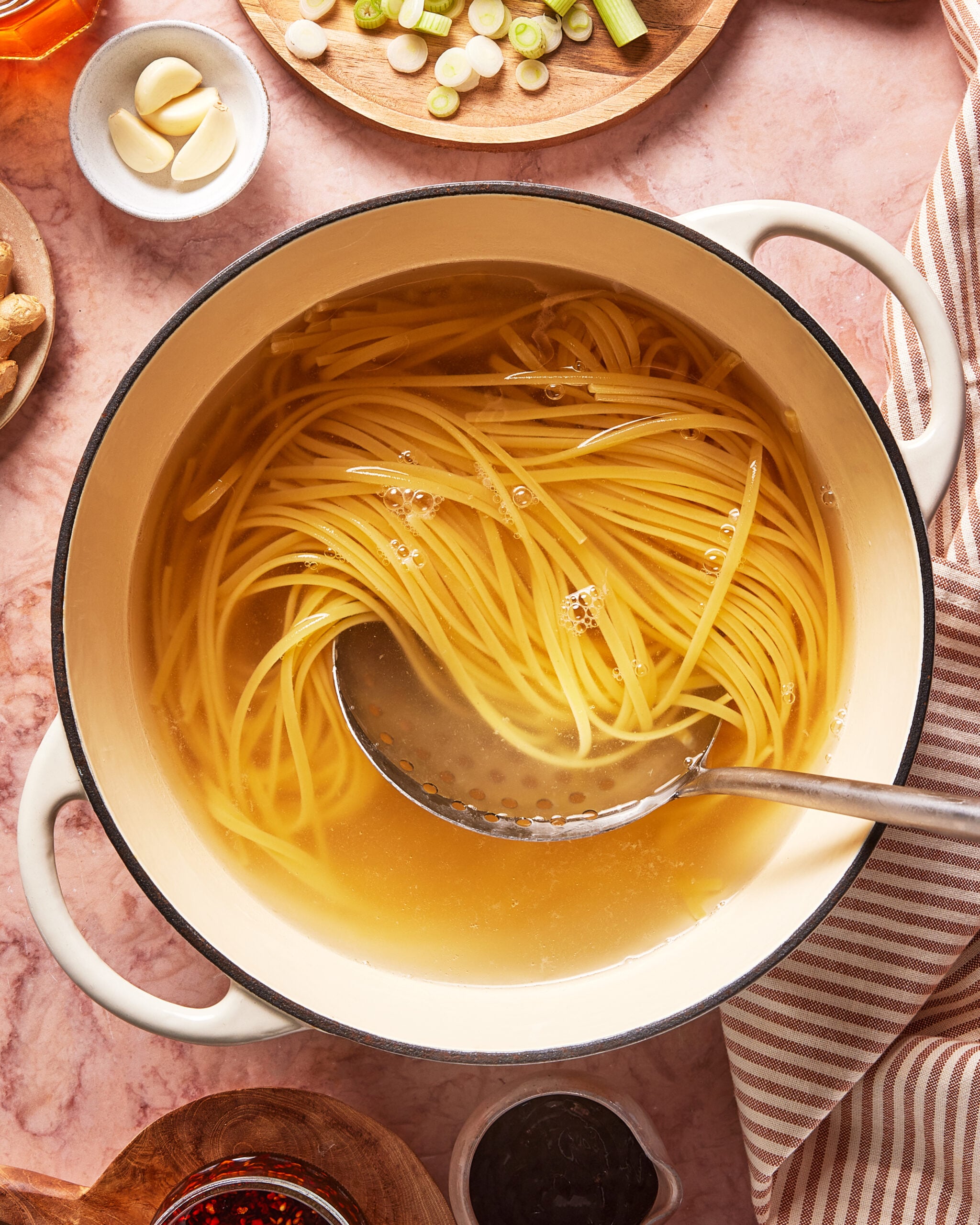A pot of spaghetti noodles cooking in clear broth, with a ladle resting inside. Surrounding the pot are ingredients such as sliced green onions, garlic cloves, and sauces on a pink countertop.