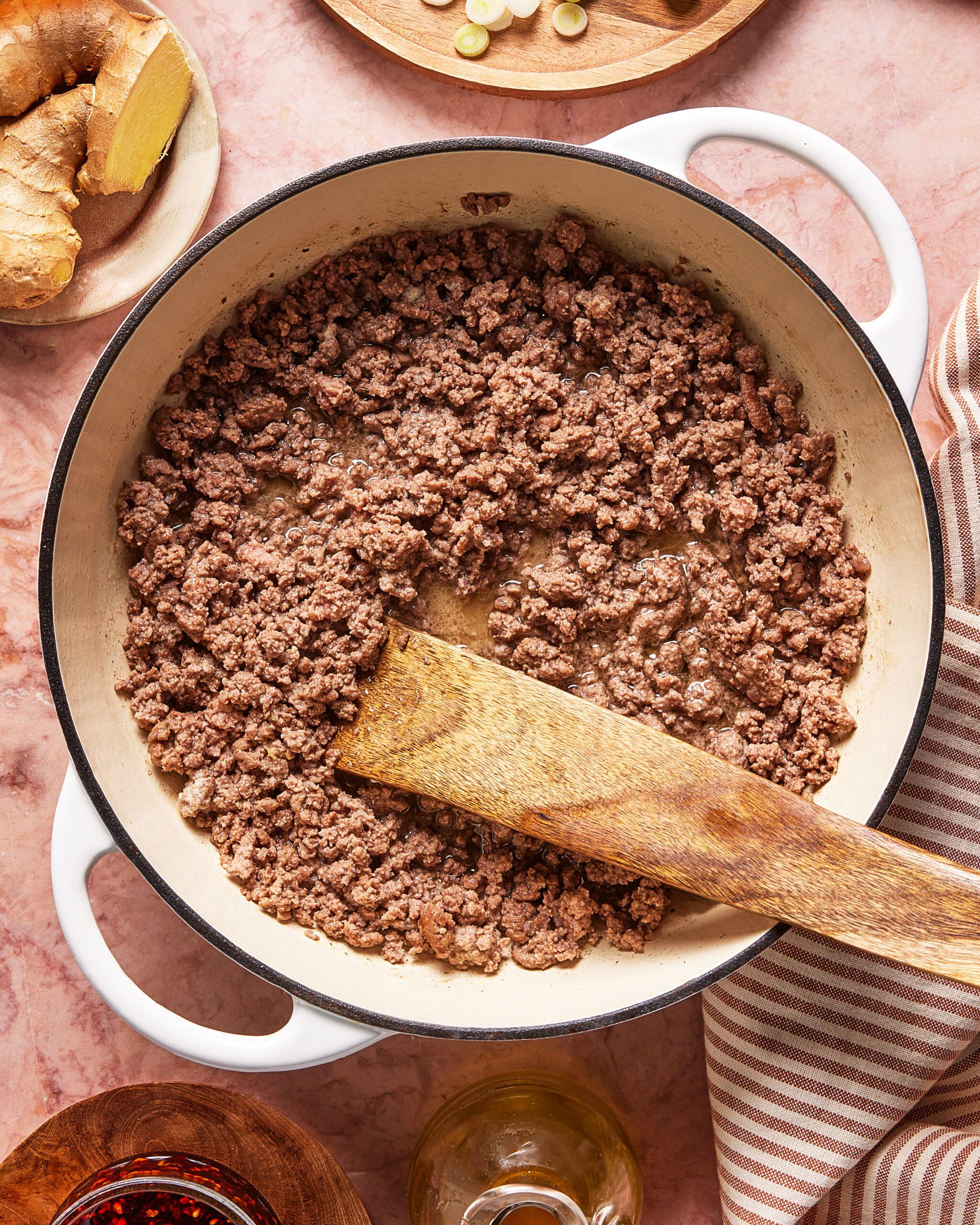 Ground beef is being browned in a white Dutch oven with a wooden spatula on a pink countertop. Ingredients like ginger, oil, and spices are arranged around the pot. A striped cloth is nearby.