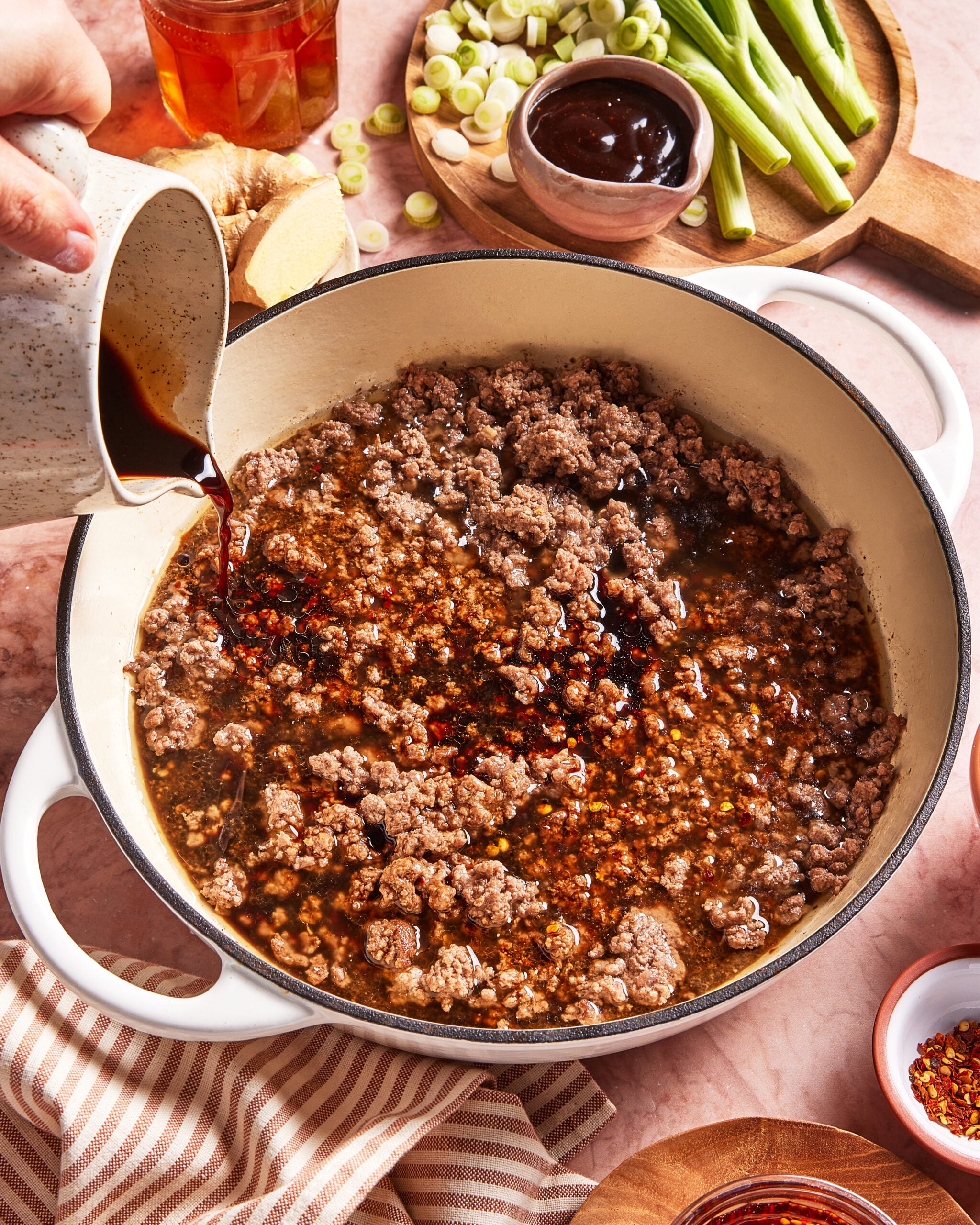 Ground meat is cooking in a white pot while a person's hand pours a dark sauce over it. Nearby are sliced green onions, hoisin sauce, ginger, a striped towel, and bowls with seasonings.
