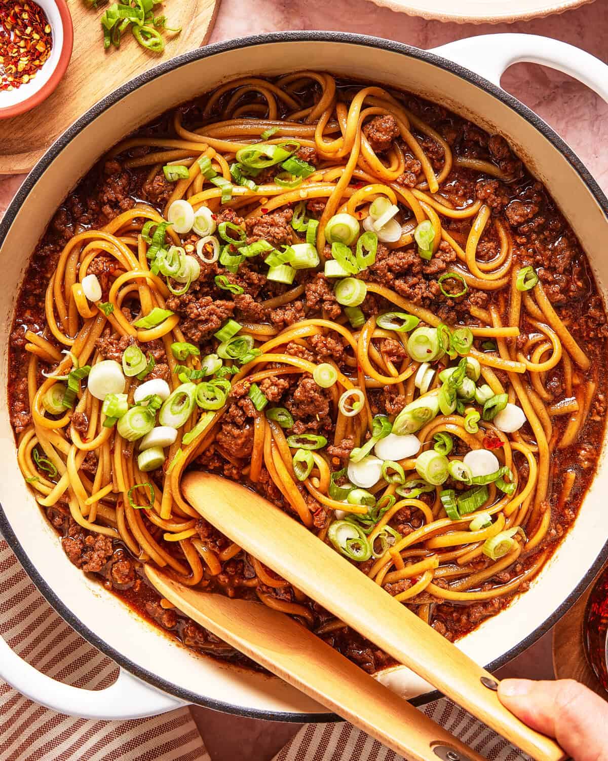 A large pot of noodles with ground meat in a rich sauce, topped with chopped green onions. Wooden tongs are mixing the dish, and a striped towel and small bowls of spices are nearby.