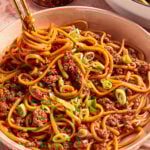 A bowl of saucy noodles mixed with ground meat and topped with chopped green onions, with chopsticks lifting a portion. A jar of chili oil and another bowl of noodles are in the background.