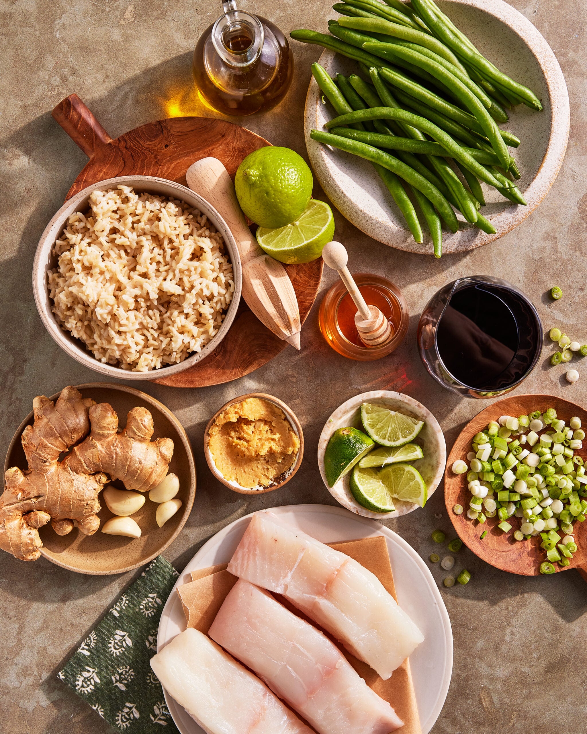 Overhead view of raw cod filets, brown rice, green beans, ginger, garlic, scallions, miso paste, lime halves, honey, olive oil, and a glass of red wine arranged on a table.