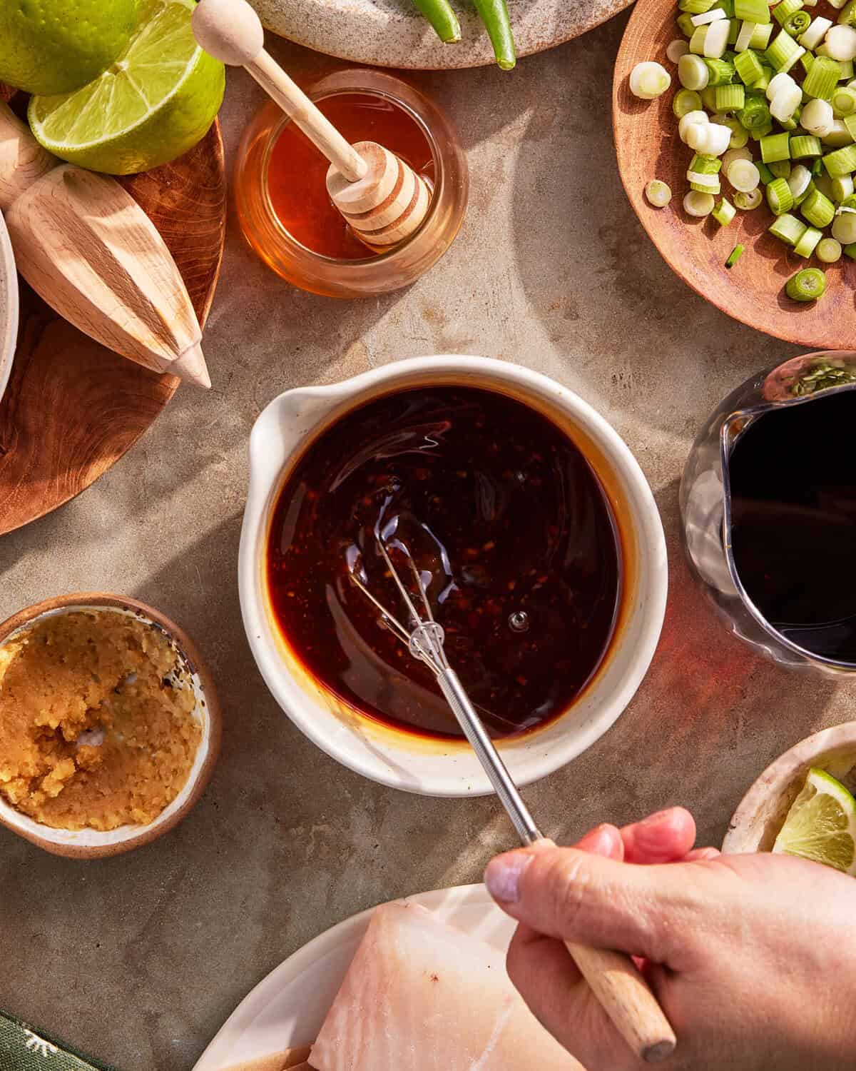 A hand holds a small whisk, stirring a dark sauce in a white bowl. Surrounding ingredients include miso paste, honey, lime, chopped scallions, green beans, and a piece of raw fish on a plate.