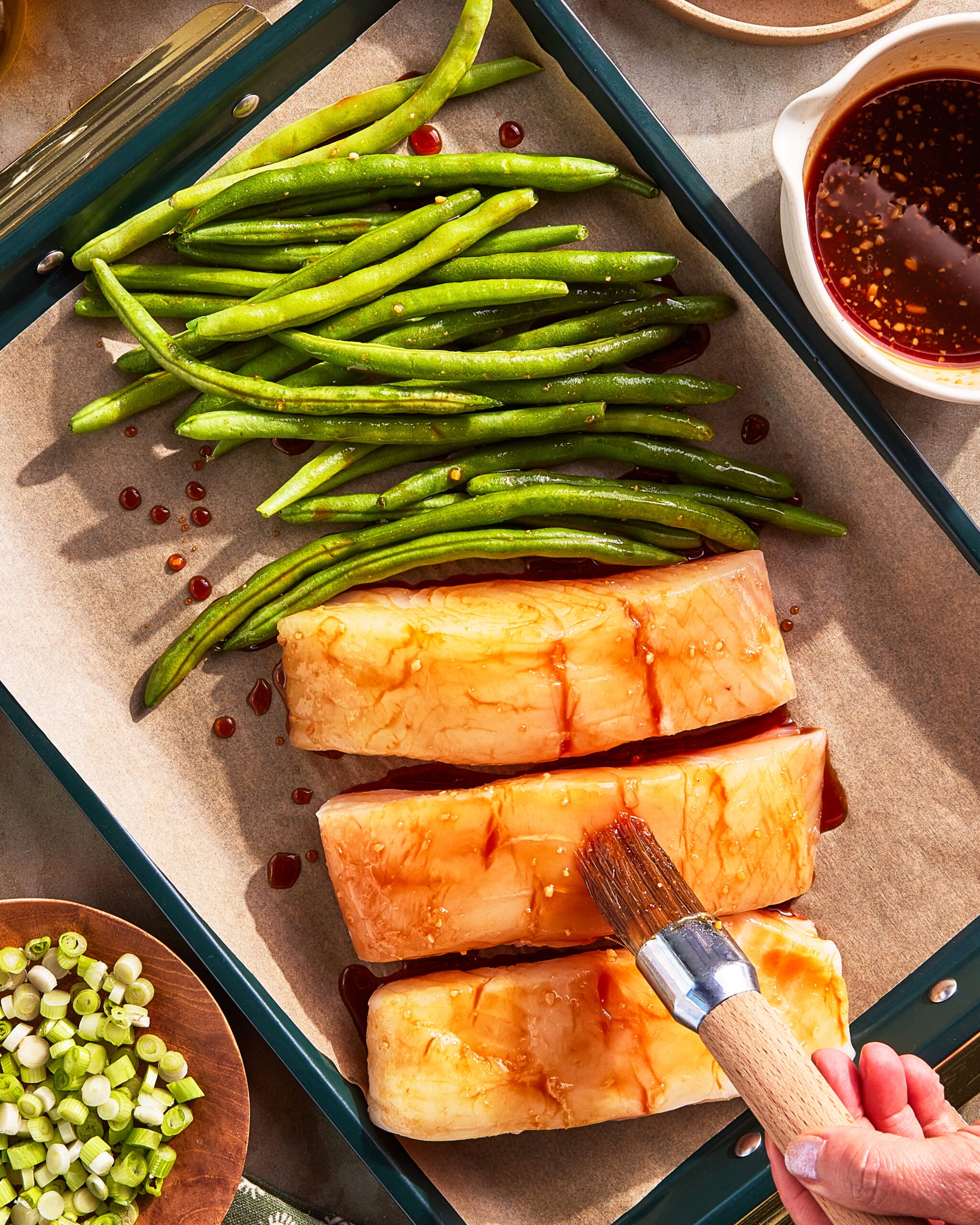 A hand brushes sauce onto tofu slabs on a parchment-lined tray beside fresh green beans. A bowl of sauce and chopped scallions are nearby, suggesting meal preparation.