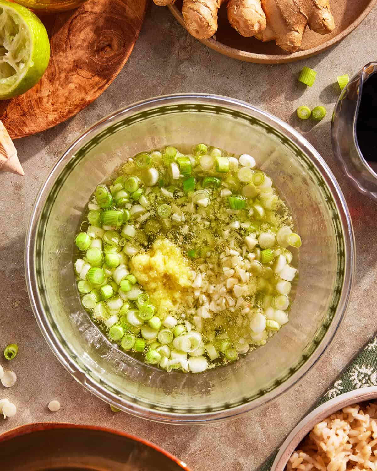 A glass bowl filled with chopped green onions, minced garlic, and grated ginger mixed with oil, surrounded by fresh ginger, a halved lime, soy sauce, and rice on a tabletop.