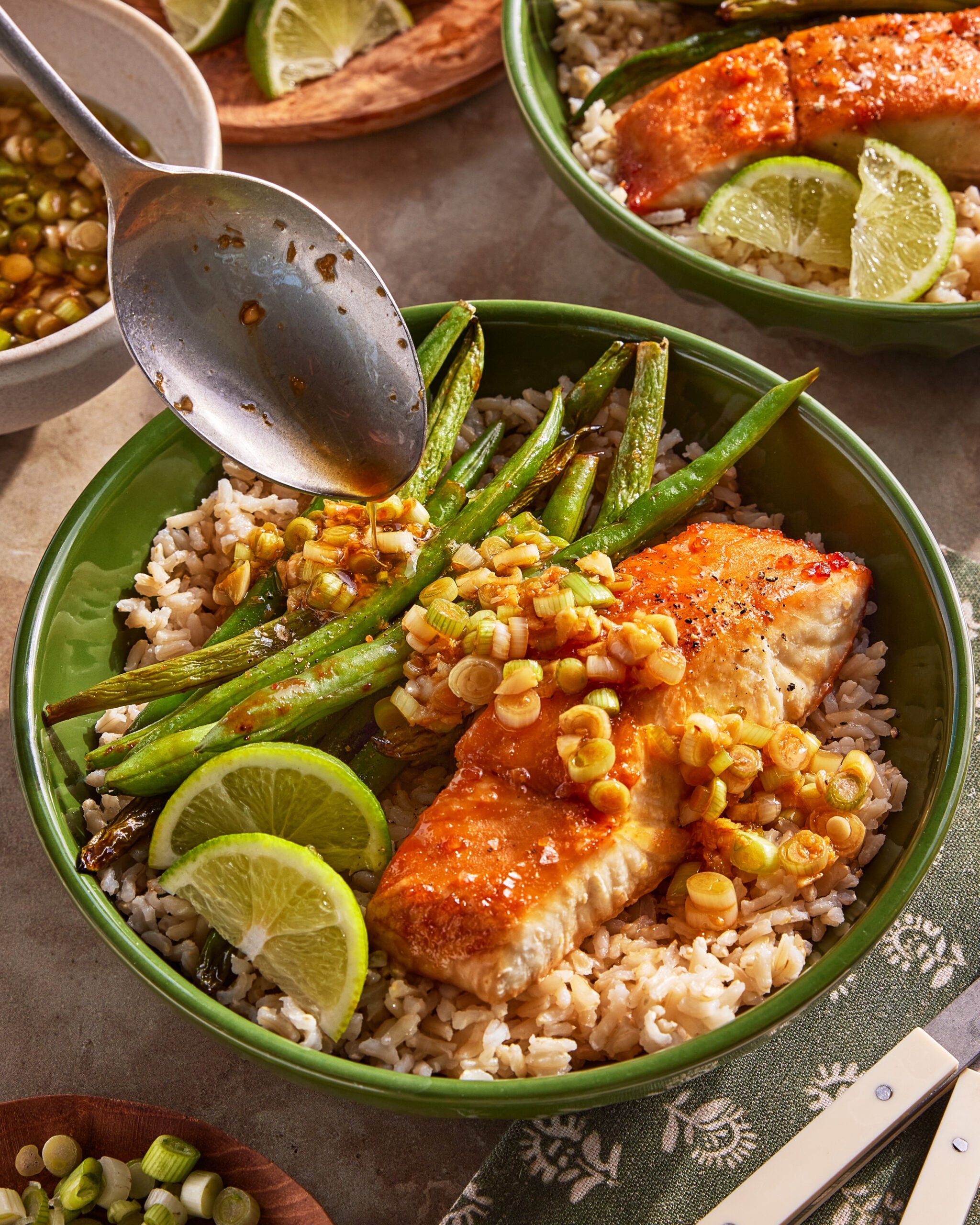 A green bowl filled with brown rice, a glazed salmon fillet, sautéed green beans, lime wedges, and chopped scallions, with a spoon drizzling sauce over the dish.