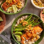 Two green bowls filled with brown rice, roasted green beans, and glazed salmon topped with sautéed scallions. Lime wedges are on the side, and a bowl of scallion sauce is nearby. Fork and knife are placed next to the bowls.