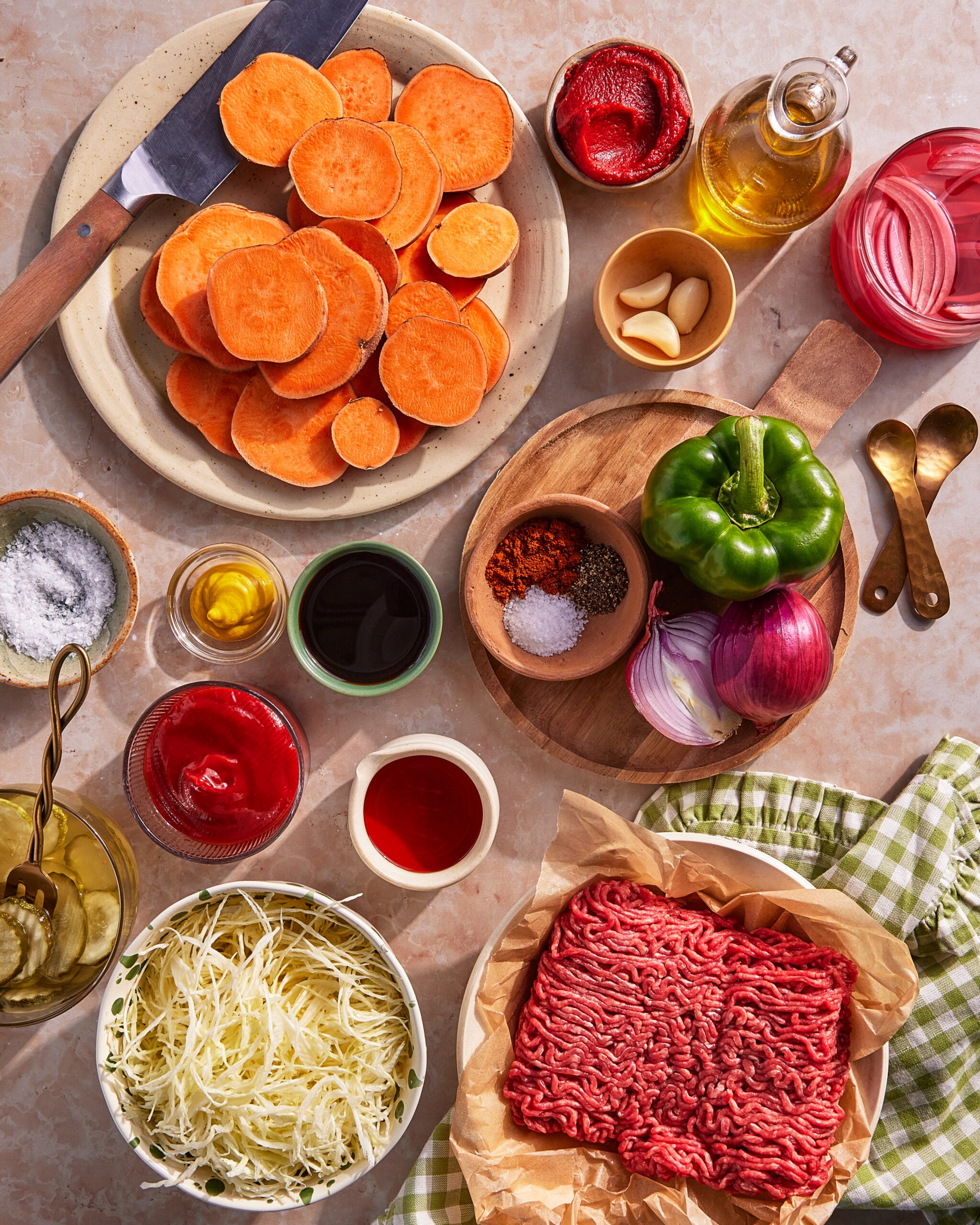 A flat lay of ingredients on a counter, including sliced sweet potatoes, ground beef, shredded cabbage, a green bell pepper, red onion, garlic, ketchup, spices, oil, vinegar, tomato paste, and salt.