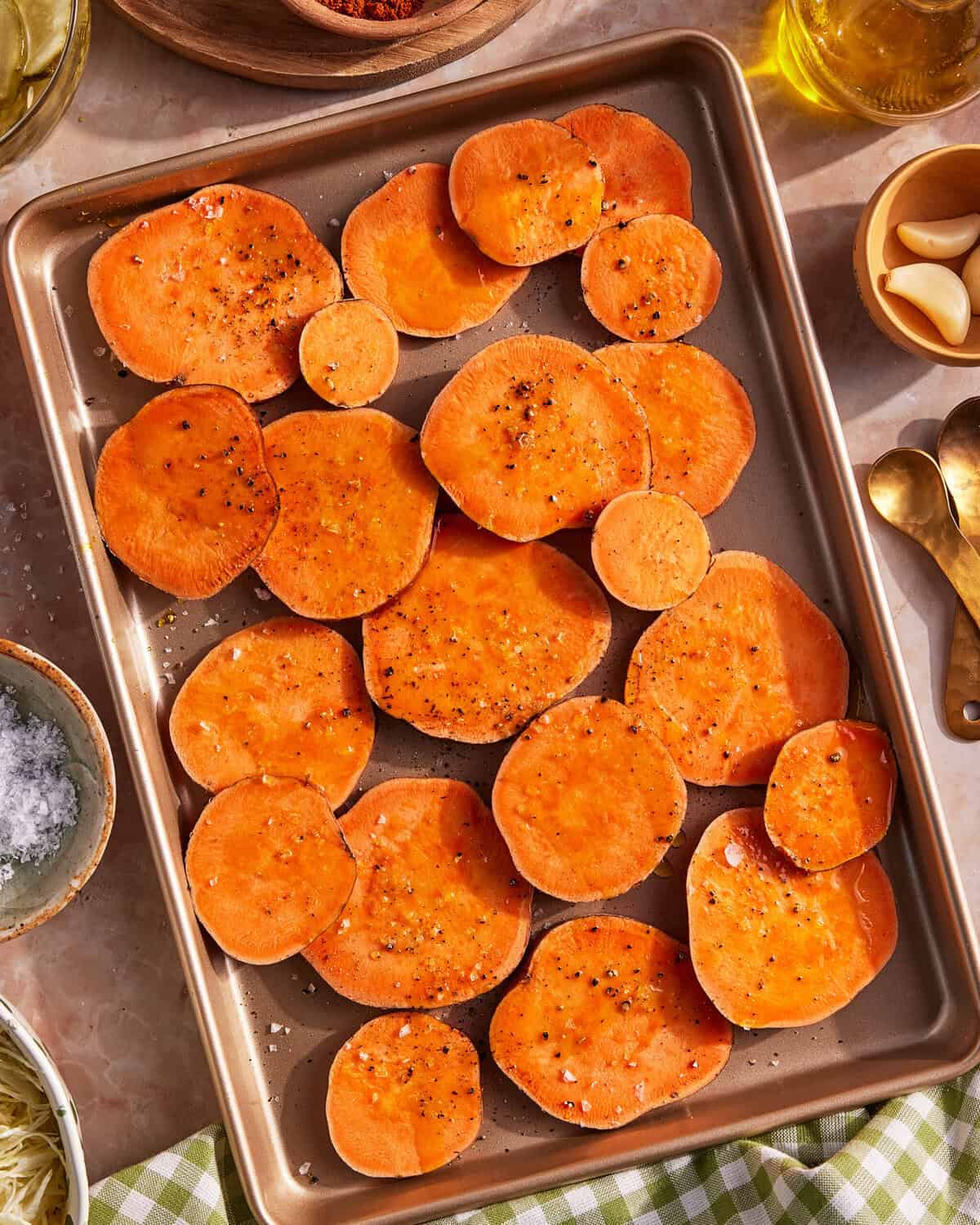 A baking tray filled with raw sweet potato slices seasoned with salt and pepper. Surrounding the tray are small bowls of salt, olive oil, garlic cloves, and gold measuring spoons on a light-colored countertop.