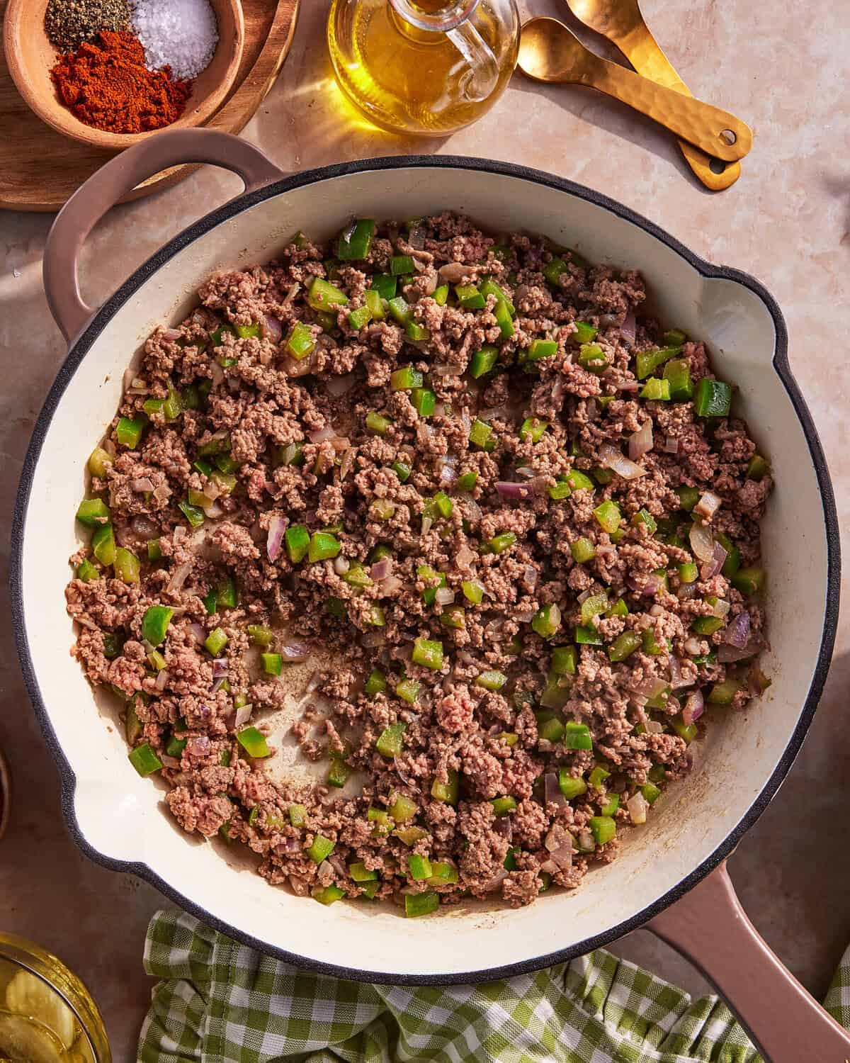 A skillet filled with cooked ground beef, chopped green bell peppers, and onions sits on a countertop next to olive oil, spices, and a green checkered cloth.