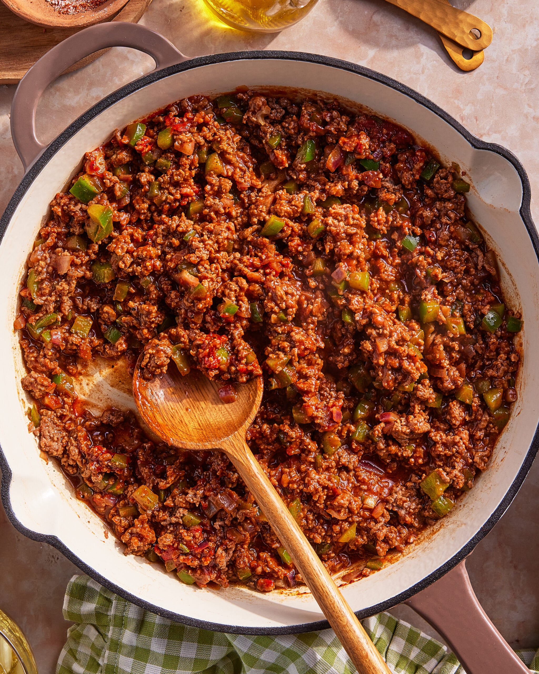 A large pan filled with cooked ground beef, diced green bell peppers, tomatoes, and onions, mixed together with a wooden spoon. The pan sits on a light surface with a green checkered cloth nearby.