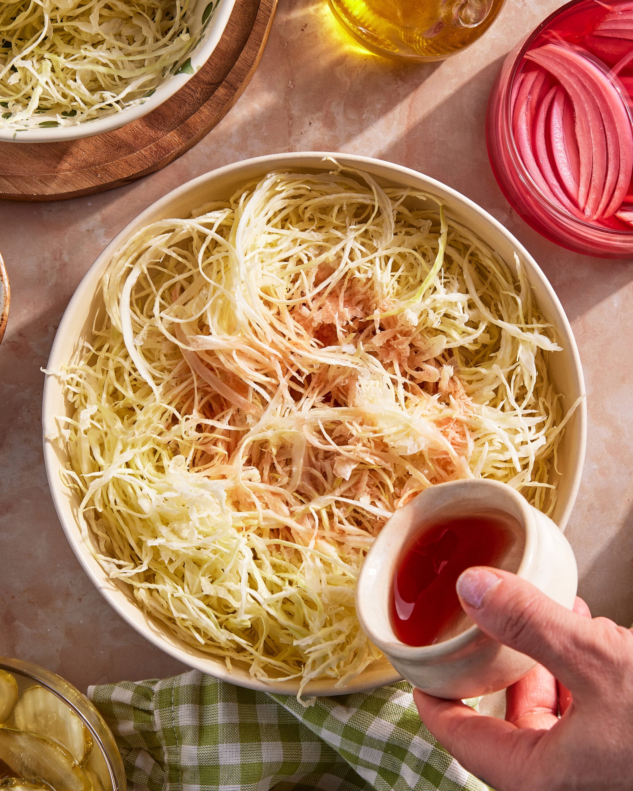 A hand is pouring red liquid, likely vinegar, from a small cup onto a bowl of shredded cabbage. Sliced onions in pink liquid, oil, and other ingredients are visible on the table.