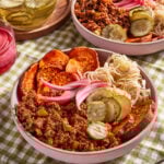 Two bowls filled with ground meat, sliced pickles, sauerkraut, roasted sweet potato slices, and pickled red onions on a green checkered tablecloth. A fork and glasses of pickles are nearby.