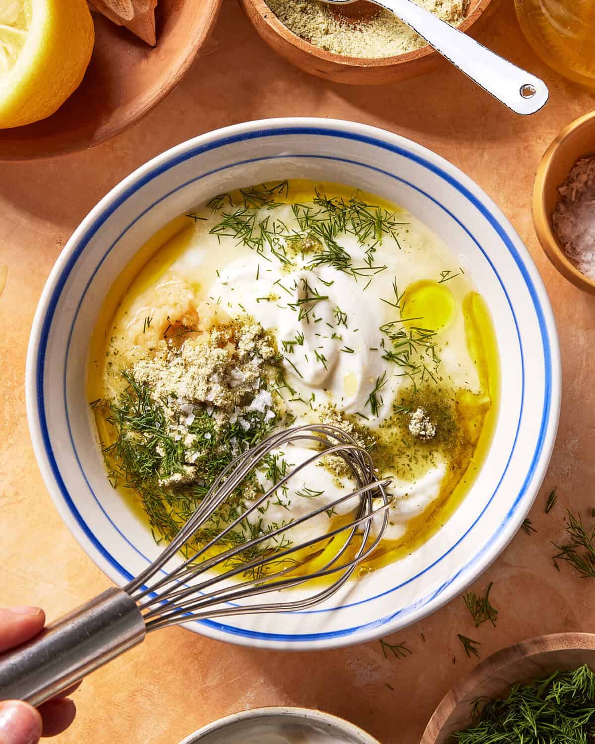 A hand holding a whisk over a bowl filled with yogurt, olive oil, herbs, and seasonings, ready to mix. Various small bowls of ingredients surround the main bowl on a tan surface.