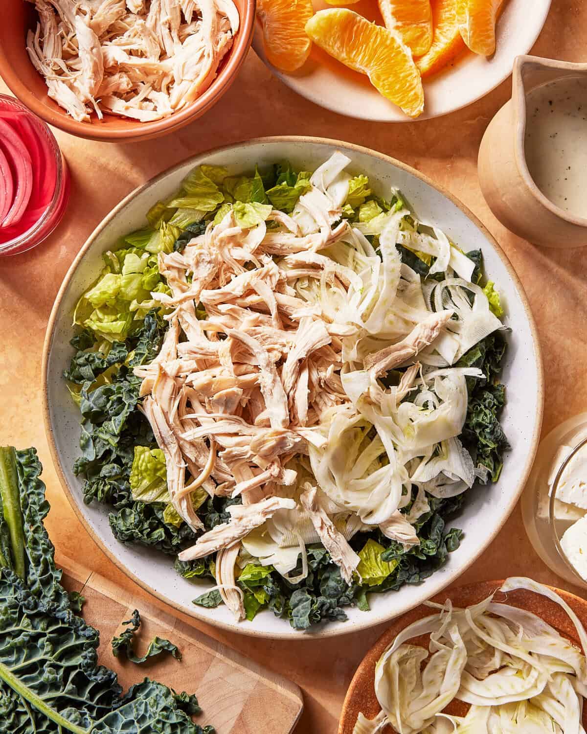 A salad bowl filled with shredded chicken, sliced fennel, and chopped leafy greens sits on a table surrounded by orange slices, dressing, feta cheese, and extra chicken in separate dishes.