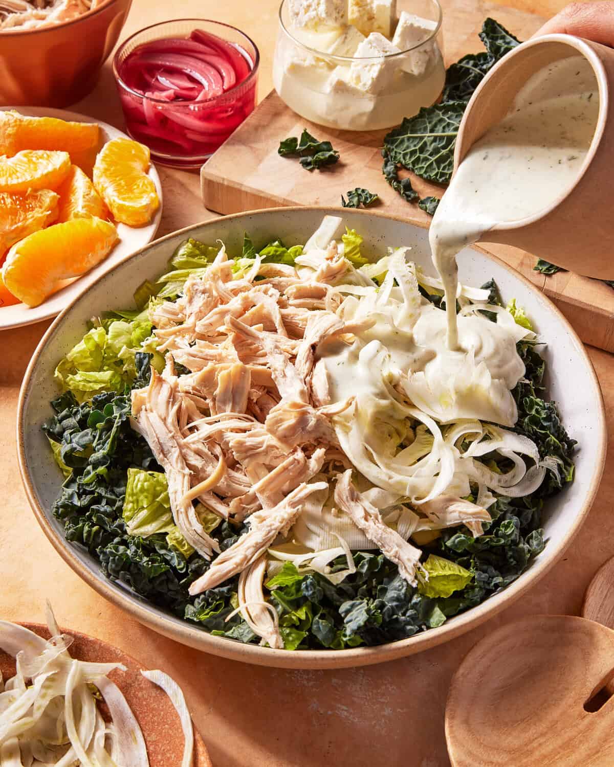 A bowl of salad with shredded chicken, sliced onions, and leafy greens is being topped with creamy dressing. Nearby are orange slices, pickled onions, feta cheese, and wooden salad servers.