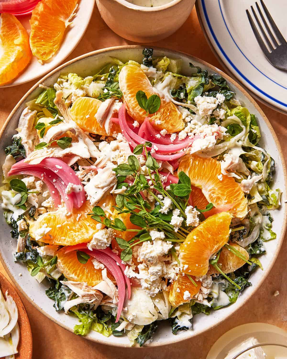 A vibrant salad with shredded chicken, orange slices, pickled red onions, crumbled feta, greens, and creamy dressing, served in a bowl on a tan surface with a plate of orange slices and utensils nearby.