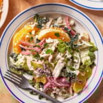 A plate of salad with shredded chicken, orange slices, pickled red onions, leafy greens, crumbled cheese, and herbs, served with a fork on a white plate with blue trim.