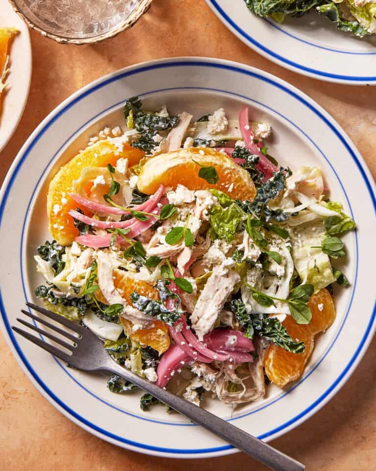 A plate of salad with shredded chicken, orange slices, pickled red onions, leafy greens, crumbled cheese, and herbs, served with a fork on a white plate with blue trim.