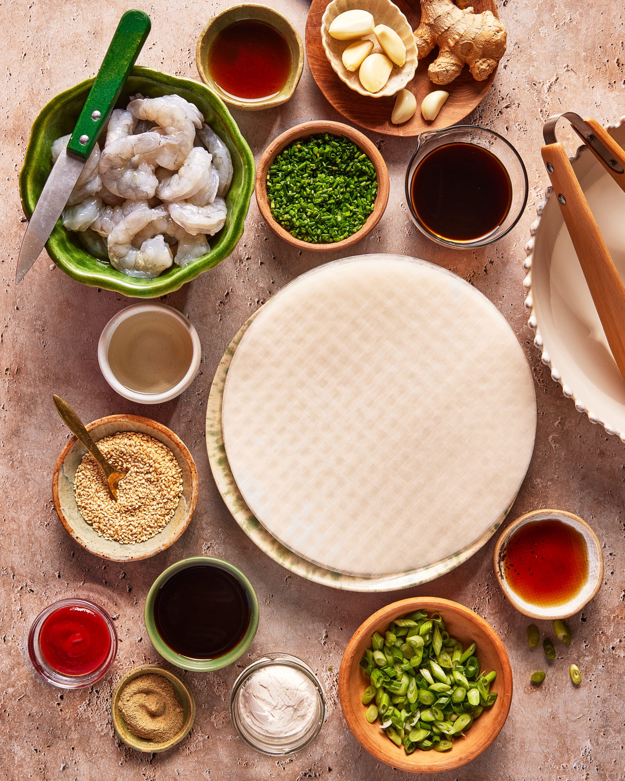 A variety of ingredients neatly arranged around a stack of rice paper wrappers, including raw shrimp, garlic, ginger, sauces, chives, sesame seeds, green onions, and a bowl of water with tongs.